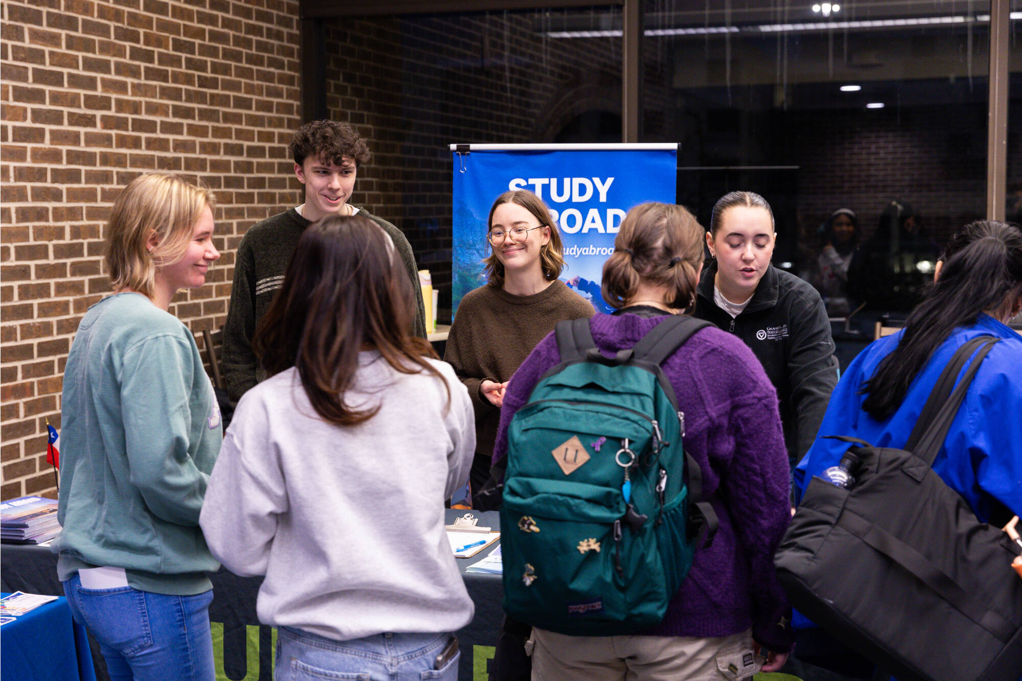 three students stand in front of a table and talk to three individuals standing behind a table.